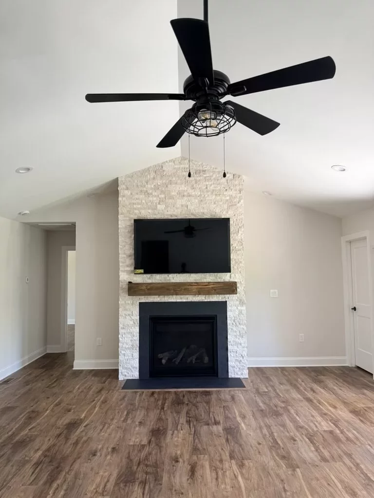 Vaulted living room with a floor-to-ceiling stacked-stone fireplace, mounted TV, and black ceiling fan