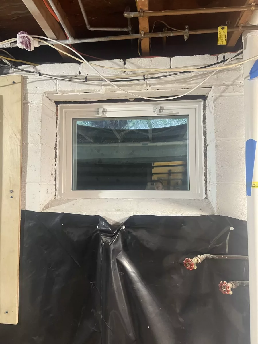 Basement window installation in progress with exposed pipes and plastic sheathing