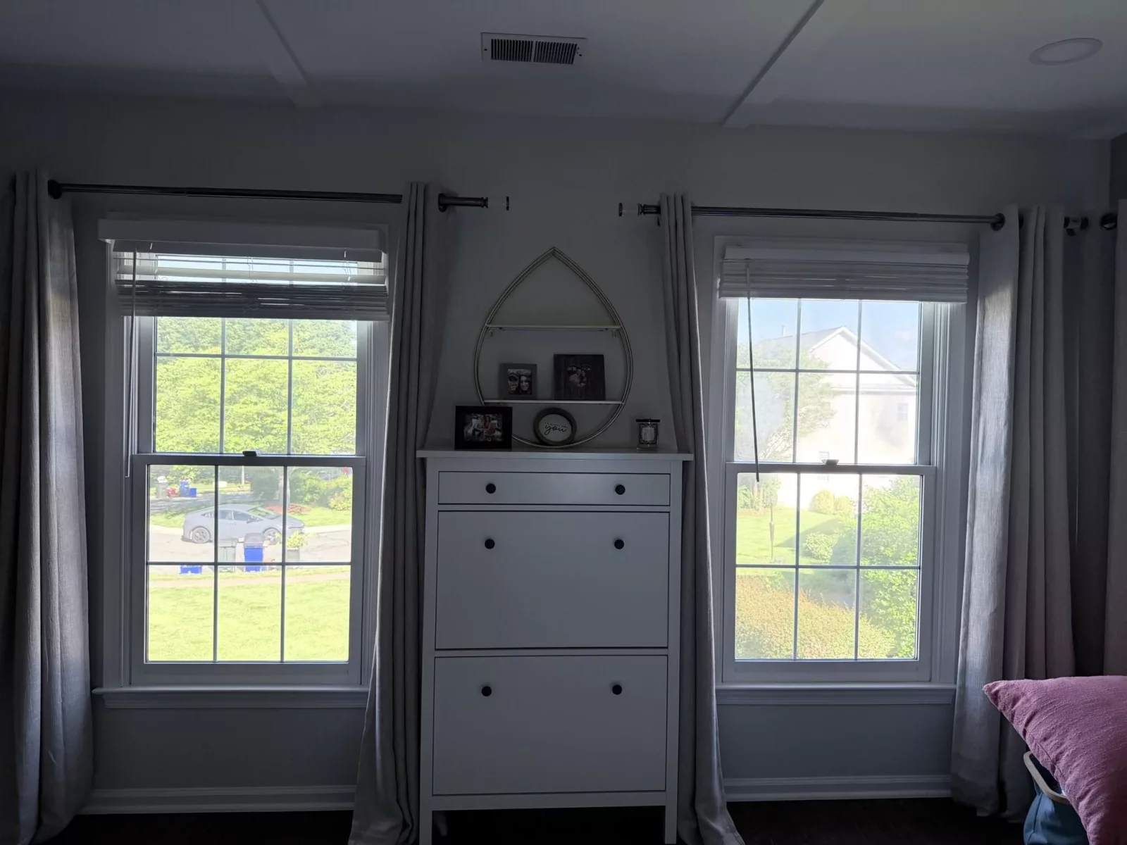 Two double-hung windows with curtains and a dresser between them in a residential room.