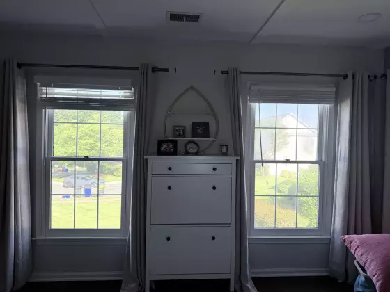 Two double-hung windows with curtains and a dresser between them in a residential room.