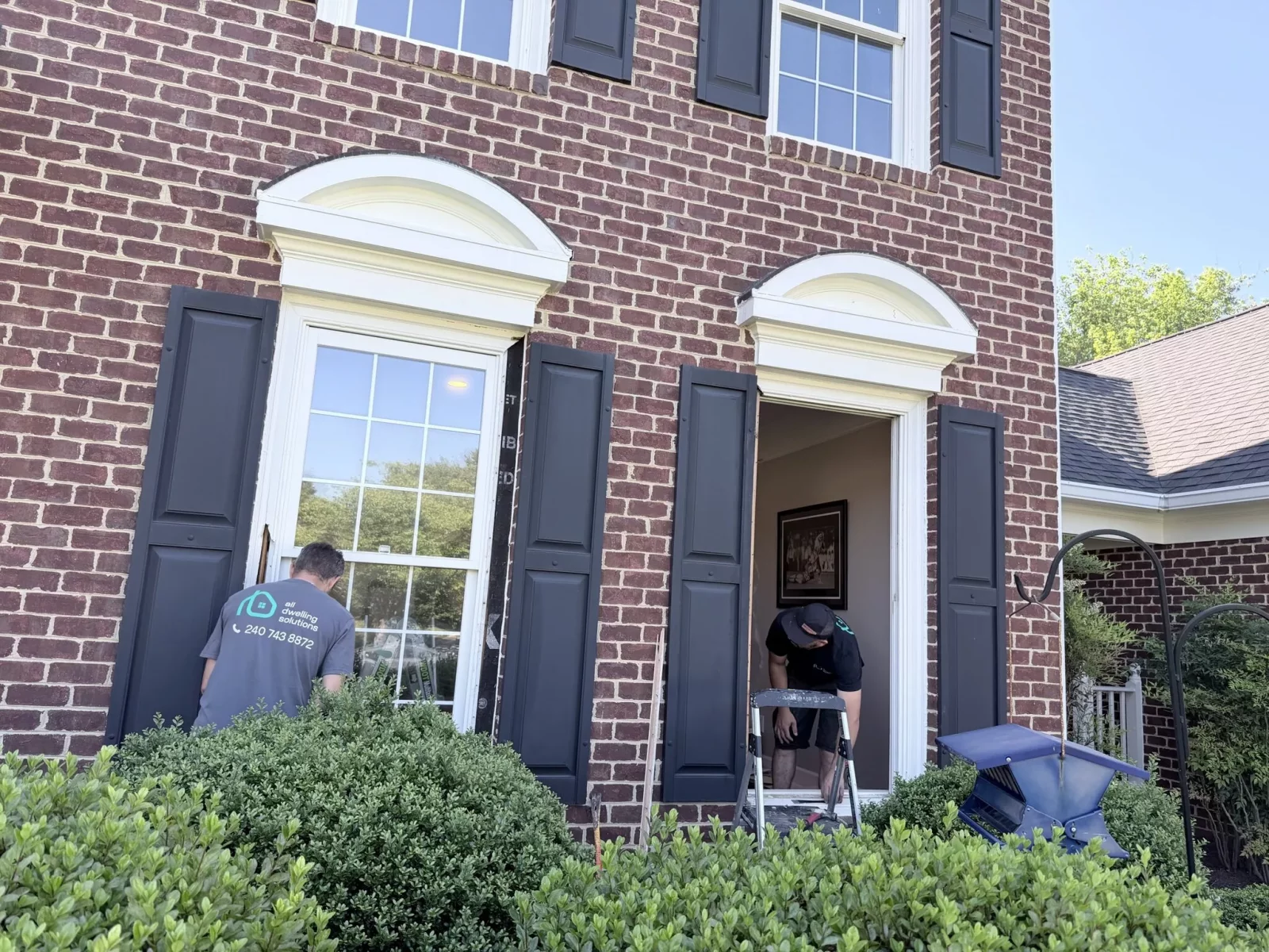 Two workers replacing windows on a brick house exterior