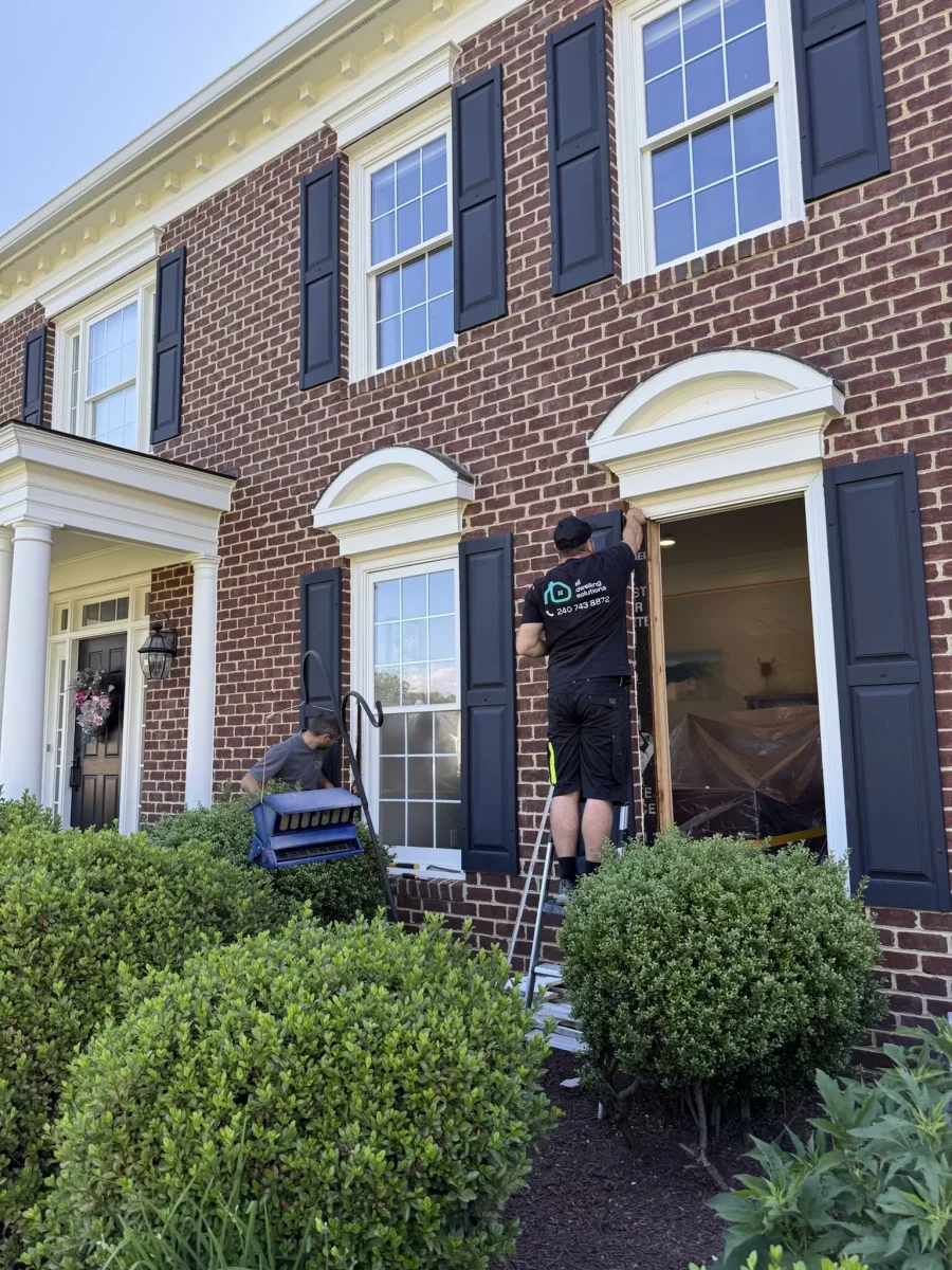 Two workers replacing a window on a brick house exterior