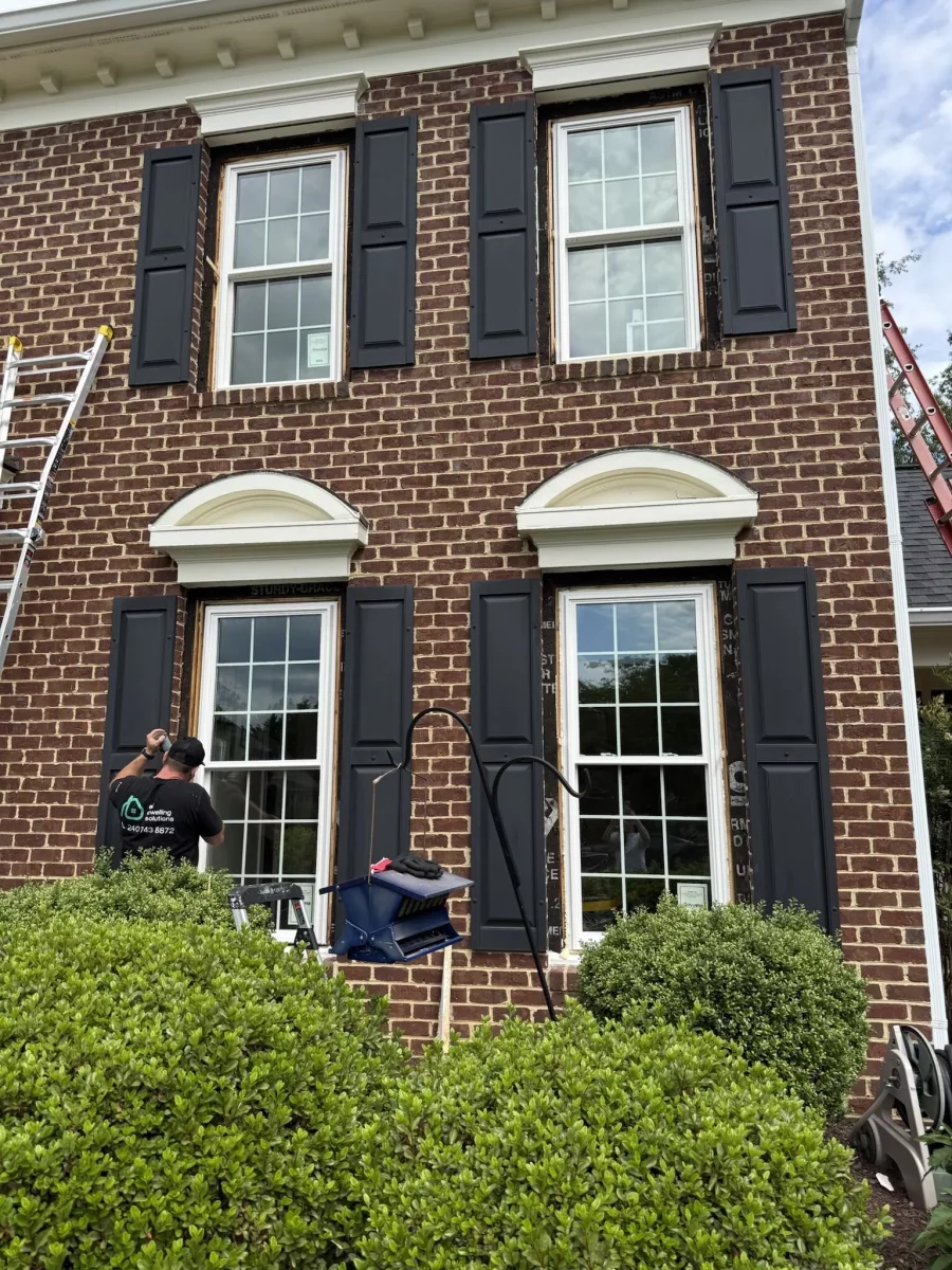 Worker installing windows on brick house exterior with shutters and ladders visible.