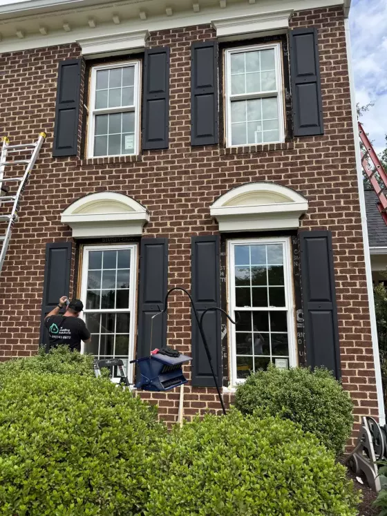 Worker installing windows on brick house exterior with shutters and ladders visible.