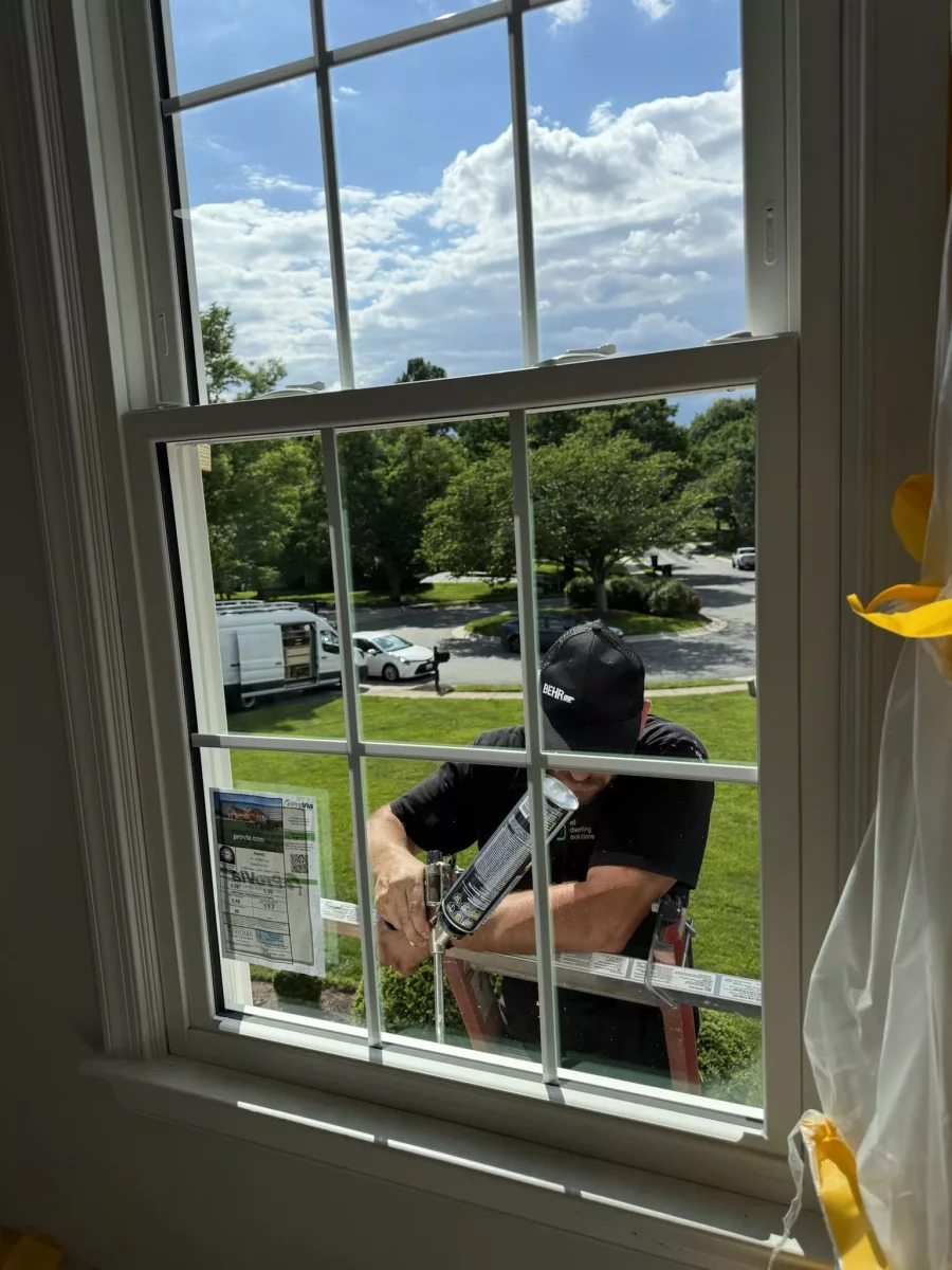Worker applying caulk to residential window from exterior ladder