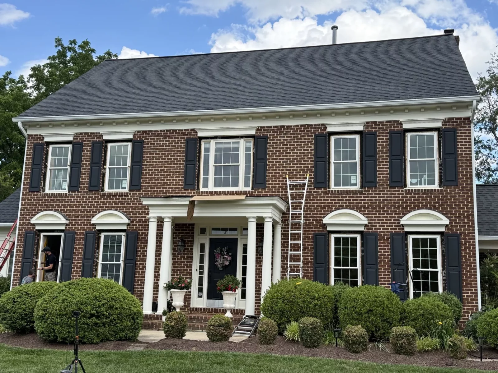 Brick house exterior with window replacement in progress, ladders and workers visible.