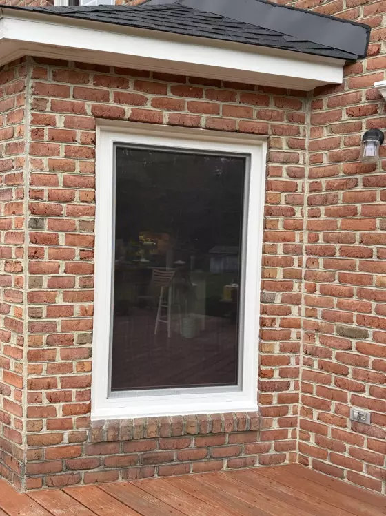 Exterior view of a single window with white trim set in a brick wall above a wooden deck.